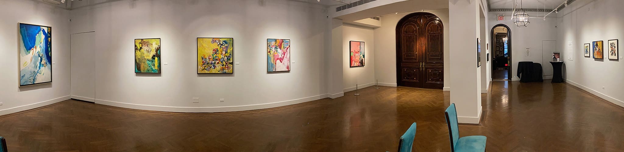 Panoramic view of an art gallery with abstract paintings and works on paper displayed on white walls, wooden floor, and a few chairs visible.