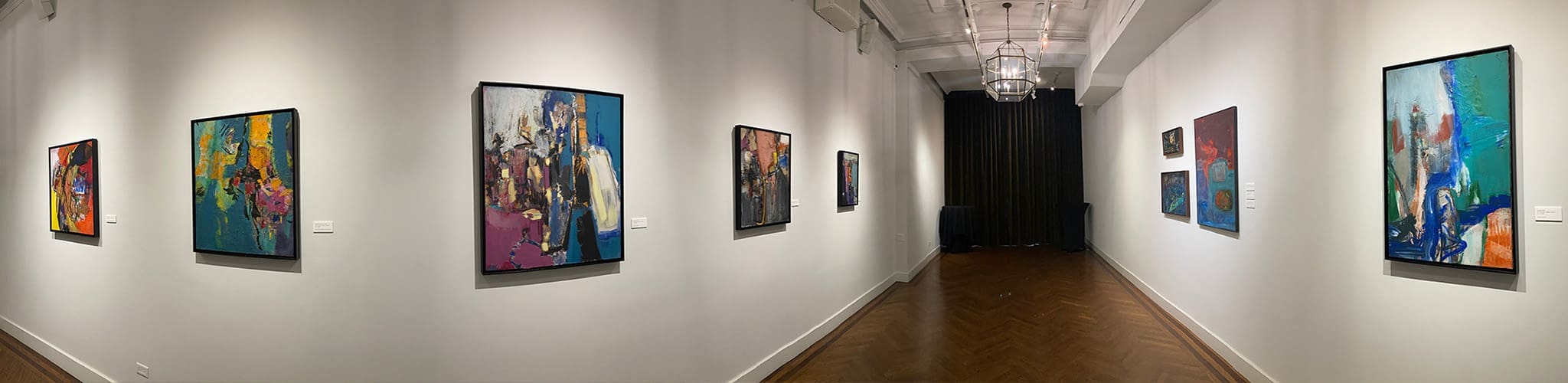 A modern art gallery with white walls displays colorful abstract paintings and works on paper in black frames; wooden floors and chandeliers complete the space.