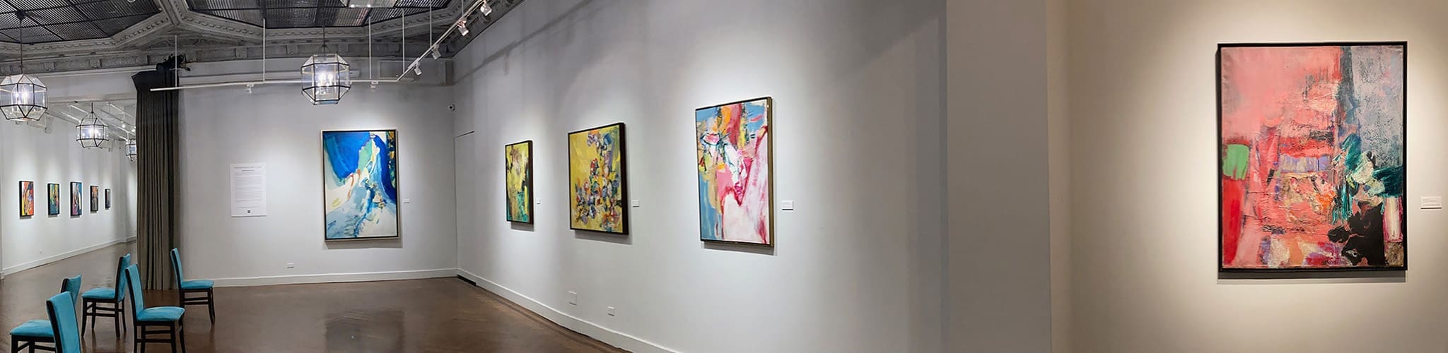 LM_2022_SOLO_Pano_04 A contemporary art gallery with abstract paintings and works on paper displayed on white walls, blue chairs lined up on the left, and overhead modern lighting.