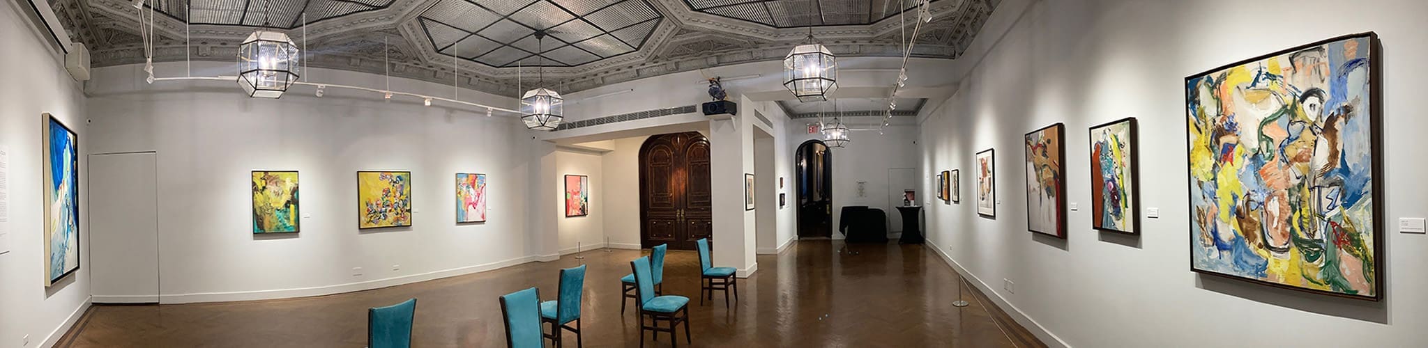 A panoramic view of an art gallery with abstract paintings and works on paper displayed on white walls, turquoise chairs arranged in the center, and ornate ceiling lighting fixtures.