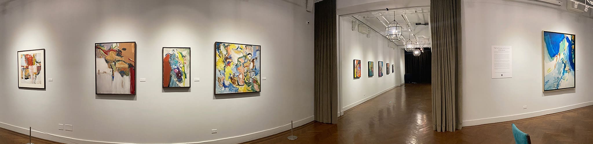 LM_2022_SOLO_Pano_06 A gallery hallway displays various abstract and colorful paintings and works on paper on white walls, with wooden flooring and overhead lighting.