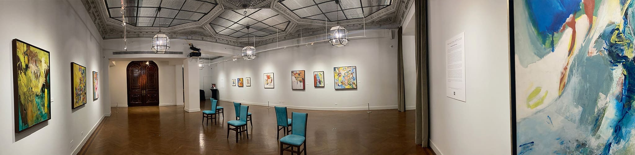 LM_2022_SOLO_Pano_07 Panoramic view of an art gallery with abstract paintings and works on paper displayed on white walls, blue chairs arranged in the center, and ornate ceiling lights overhead.